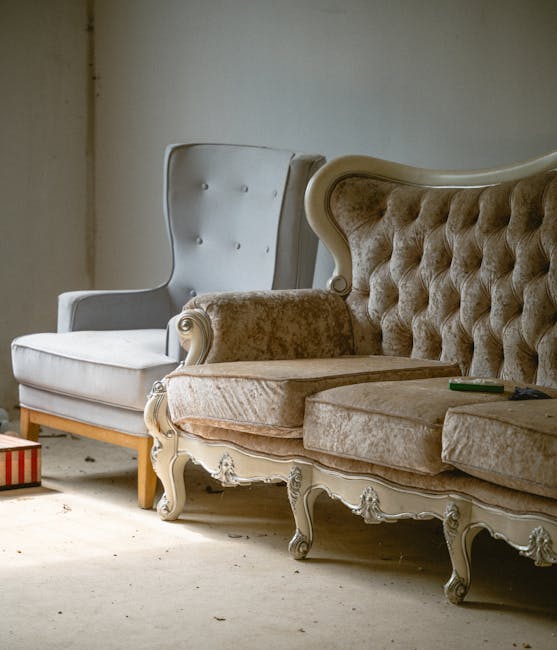 The image shows two vintage armchairs placed inside a room, likely for a house removal or furniture clearance. The first armchair, positioned on the left, has a white upholstery with button-tufted backrest and wooden legs, while the second, on the right, features a beige, velvet-like fabric with a high, curved back and ornate carved wooden legs. The beige armchair appears to be partially disassembled or prepared for moving, with some fabric cushions removed or loosely placed on top. The floor is light-colored, possibly wood or laminate, and the background wall is plain and light-colored, with visible dirt or marks near the baseboard. There are no other items visible in the room. This scene suggests a furniture collection or packing process associated with a house relocation, where [COMPANY_NAME] may be coordinating the careful loading of these chairs—along with similar items—into a vehicle for transport, highlighting the importance of professional removals, packing, and logistics during home moves. The environment is well-lit, likely by natural light, emphasizing the furniture's condition and the preparatory state for moving.