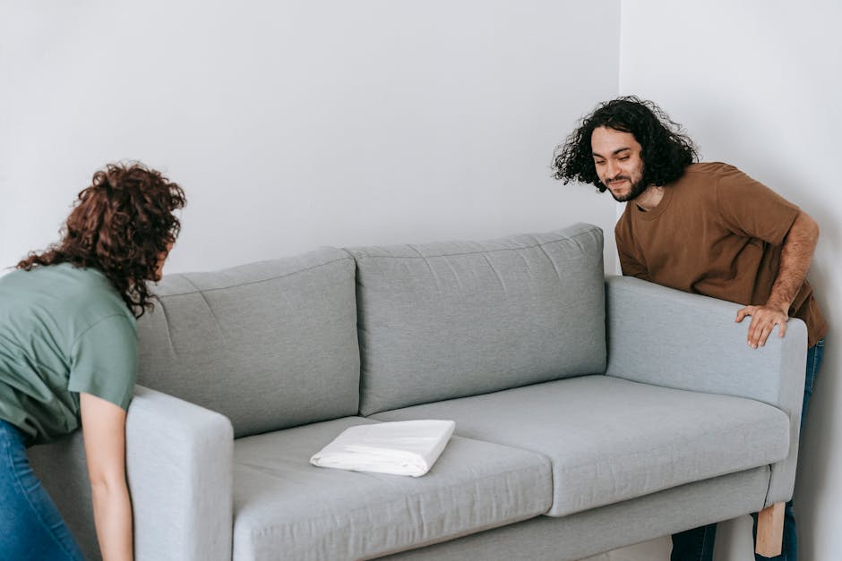 A man and a woman are performing a home relocation task indoors, lifting a light grey fabric-upholstered sofa with wooden legs. The woman, wearing a green t-shirt and jeans, is positioned on the left side, slightly bent forward gripping the armrest, while the man, dressed in a brown t-shirt and dark trousers, stands on the right side holding the opposite armrest. Both are focused on carefully moving the sofa across a room with plain white walls. On the sofa, there are some white packing blankets or cushions neatly folded, indicating preparation for safe transportation. In the background, there are no visible doors or windows, but the area appears to be inside a residential property, consistent with furniture transport and packing during a house move. The scene suggests a coordinated effort as part of a professional removals service, such as those offered by Man with Van Deptford, to facilitate furniture transport and home relocation processes.
