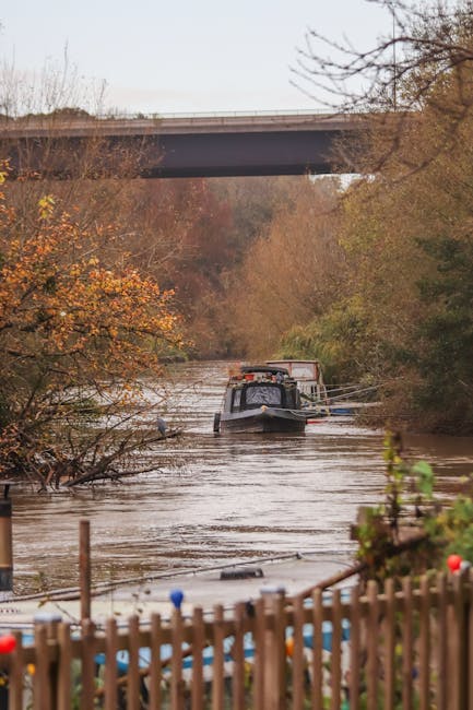 Image showing a narrow river or canal surrounded by trees with autumn foliage, featuring a small boat or barge being navigated through the water. The boat is positioned centrally in the image, with visible details of its structure, including a cabin area and some cargo or equipment on deck. In the background, a concrete bridge spans across the river, partially obscured by the tree branches. At the water's edge, a wooden fence runs along the foreground, with some floating debris or objects visible near the surface. The scene appears natural and outdoors, with muted lighting indicative of an overcast day. This setting aligns with the context of transportation and logistics, similar to the processes involved in home relocations or furniture transport with [COMPANY_NAME], supporting the theme of moving services associated with the page title, 'Creek Road to Deptford Market Yard: navigating tight removals, DEPTFORD.'