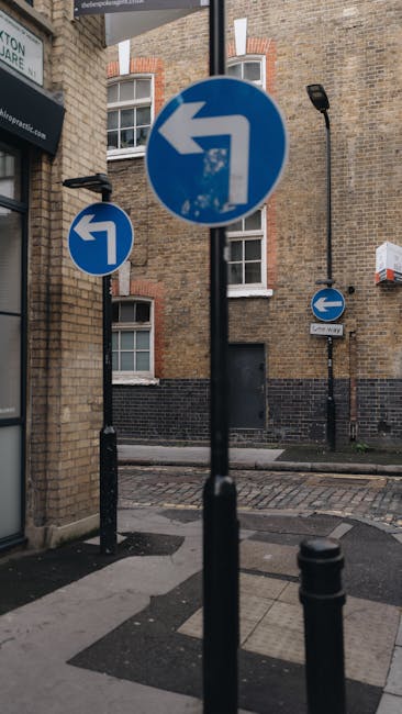 The image shows a street corner with three blue circular traffic signs indicating left turns, mounted on black poles. The signs are positioned against a backdrop of a multi-storey brick building with white-framed windows and a black door, typical of an urban residential or commercial area in Deptford. A black lamppost is also visible on the sidewalk nearby, along with a street sign labeled 'One Way,' indicating the direction of traffic flow. The pavement consists of grey concrete slabs, and a small section of cobblestone paving can be seen at the street intersection. The scene is well-lit with natural daylight, and the overall setting suggests an environment suitable for home relocation or furniture transport activities, which [COMPANY_NAME] might conduct as part of their removals services, emphasizing safe loading and navigation through urban areas.