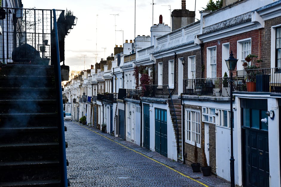 A row of terraced houses in a narrow street during daylight, featuring brick facades, small front gardens with potted plants, white window frames, and external staircase access to upper floors. The pa