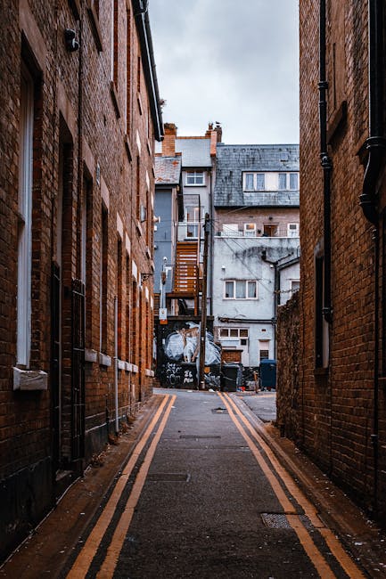 A row of terraced houses in a narrow street during daylight, featuring brick facades, small front gardens with potted plants, white window frames, and external staircase access to upper floors. The pa