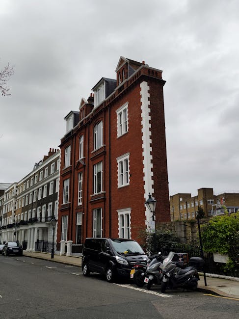 Photograph of a narrow Victorian-style red brick townhouse with white window frames and decorative white vertical stripes along the side. The building is situated on a city street under an overcast sky, with other similar terraced houses in the background. In front, there are parked vehicles including a black van and multiple motorcycles or scooters on the pavement, positioned near the entrance. The street appears quiet, with a lamp post and some greenery visible. This setting may relate to house removals or home relocation services in Deptford, where detailed planning and careful transportation of furniture and belongings are essential, as handled by Man with Van Deptford.