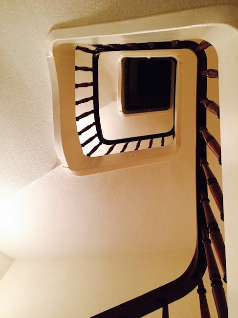 A top-down view of a narrow Victorian staircase inside a property, showing wooden steps with a weathered appearance, metal handrails on either side, and a cylindrical concrete column at the top of the stairs. The staircase is enclosed by walls with a light grey finish, and natural light illuminates the area, highlighting the textures of the wood and concrete. The image captures the confined space typical of heritage homes in Deptford. This setting is part of a home relocation process managed by Man with Van Deptford, illustrating the careful planning needed for furniture transport and packing within tight stairwell layouts.