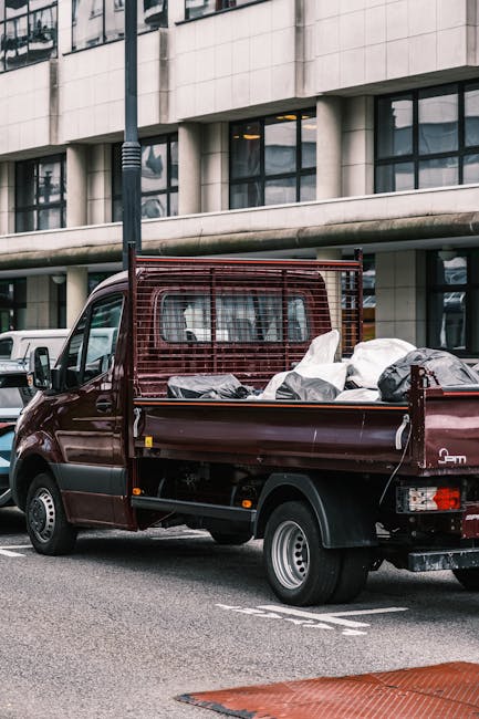 A dark red flatbed van parked on a city street in front of a modern commercial building with large windows and an overhanging concrete ledge. The van's cargo area is filled with various household item
