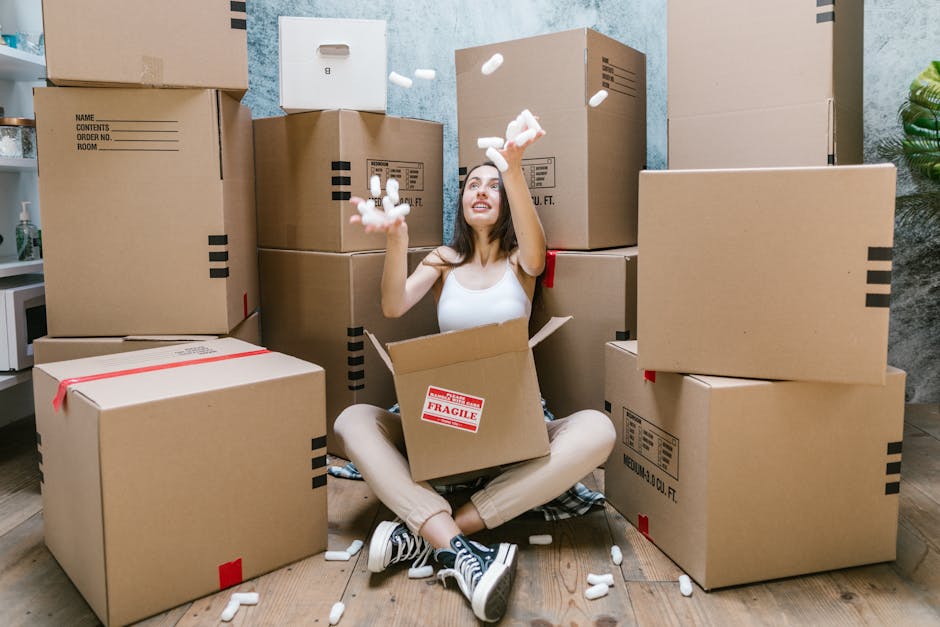 A young woman is seated on a wooden floor inside a room, surrounded by numerous large cardboard moving boxes of varying sizes, some with red fragile stickers and black markings indicating contents. She is smiling and looking up as she throws white packing peanuts into the air above her hands. The packing materials are scattered around her, with some spilling out of her open box that is positioned on her lap. The room has a light textured wall in the background, and a small potted plant can be seen on the right side. The boxes are stacked against the wall, with a doorway partially visible to the left, suggesting an indoor home environment suitable for furniture transport and home relocation activities. This scene reflects the packing and moving process involved in a house removal, highlighting the careful handling and organization necessary for an efficient relocation by a professional service such as Man with Van Deptford.