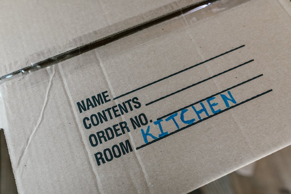 A cardboard moving box labeled with a printed and handwritten section indicating contents, with the words 'NAME', 'CONTENTS', 'ORDER NO.', and 'ROOM', where 'KITCHEN' is handwritten in blue ink. The box is made of brown corrugated cardboard with visible tape along the top edges. It is placed inside a property, possibly near an entrance or hallway, with some plastic wrapping visible around it. The environment appears well-lit, and the box is part of a home relocation or furniture transport process managed by Man with Van Deptford, which specializes in professional removals and packing services. The image captures the packing stage of an emergency or late-notice move, focusing on organized packing materials prepared for loading onto a moving van, with the box situated on the floor, ready to be carried or loaded into the vehicle for transport to the new address.