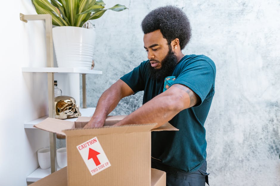 A young woman is seated on a wooden floor inside a room, surrounded by numerous large cardboard moving boxes of varying sizes, some with red fragile stickers and black markings indicating contents. She is smiling and looking up as she throws white packing peanuts into the air above her hands. The packing materials are scattered around her, with some spilling out of her open box that is positioned on her lap. The room has a light textured wall in the background, and a small potted plant can be seen on the right side. The boxes are stacked against the wall, with a doorway partially visible to the left, suggesting an indoor home environment suitable for furniture transport and home relocation activities. This scene reflects the packing and moving process involved in a house removal, highlighting the careful handling and organization necessary for an efficient relocation by a professional service such as Man with Van Deptford.