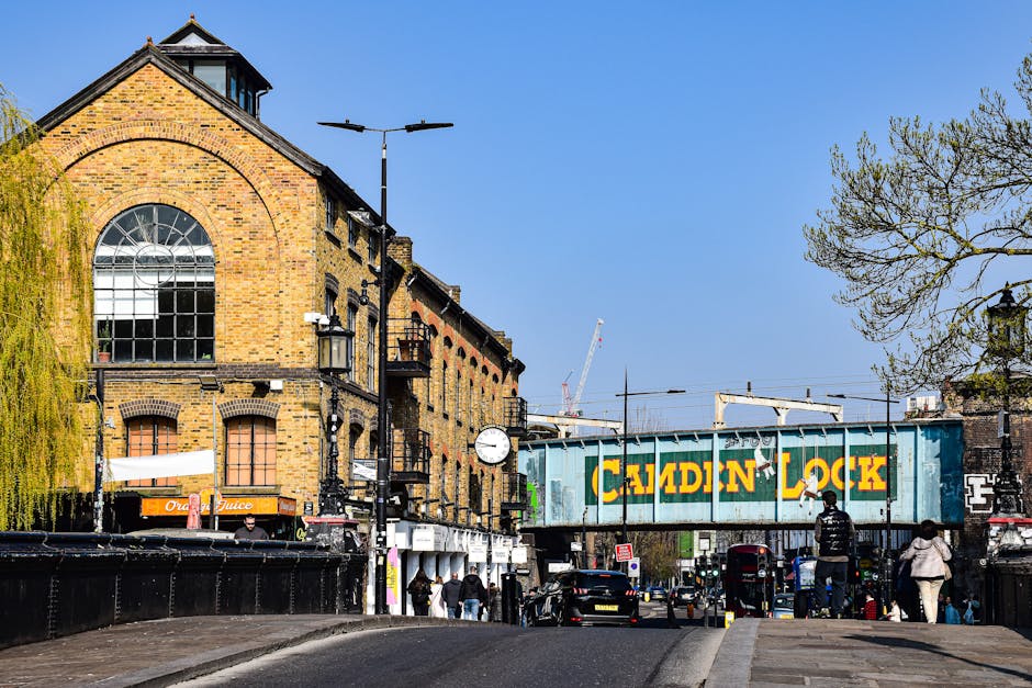 A white flatbed truck from Man with Van Deptford is parked on a city street in Lewisham, loaded with various furniture items including wooden pallets, cardboard boxes, and wrapped packages, ready for