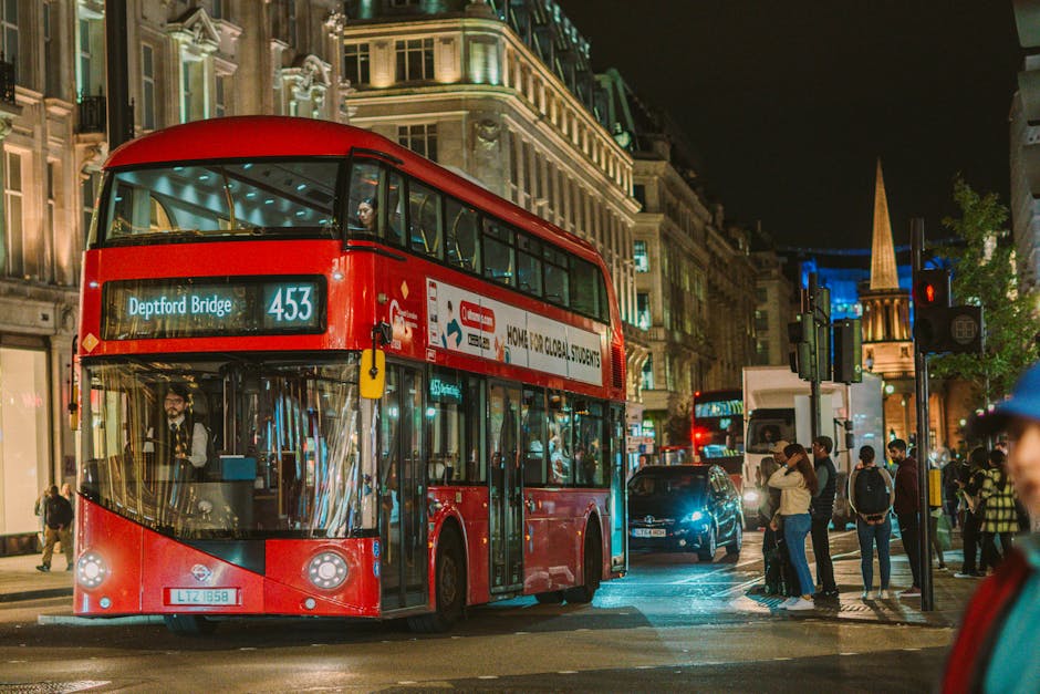 A double-decker red bus numbered 453 displaying 'Deptford Bridge' as its route destination is stationary at a busy street corner during nighttime. The bus is illuminated, with visible windows revealing seated passengers inside. Pedestrians are waiting at the bus stop or crossing the road, some carrying backpacks, wearing jackets and hats suitable for cool weather. The street scene includes various vehicles, such as a car with headlights on, and nearby buildings with illuminated facades and windows. In the background, a church spire rises above the cityscape, with streetlights and traffic signals contributing to the vibrant urban atmosphere. This scene may be associated with streets in Deptford, illustrating the typical environment for house removals or moving services in the area. Man with Van Deptford occasionally facilitates such urban relocations, often involving packing, loading, and transport processes in busy city streets.