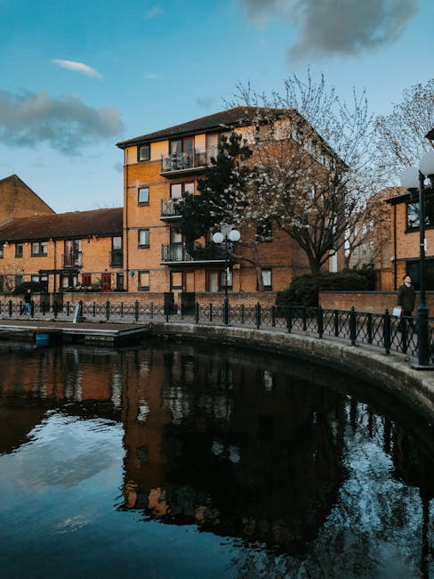 A multi-storey brick residential building located on Surrey Canal Road in Deptford, with small balconies and large windows, situated behind a stone and metal fence along a canal or riverbank. In the foreground, there is a paved walkway with street lamps and a few pedestrians, some of whom are walking or standing still. A large, leafless tree is positioned near the building, partly obscuring the view of the lower floors. The water in the canal reflects the sky, trees, and part of the building, indicating calm water conditions. The scene is captured in daylight with a partly cloudy sky, showing early evening lighting. This setting illustrates a typical urban environment suitable for house removals and furniture transport, where [COMPANY_NAME] might facilitate home relocation, loading, and unloading processes around such residential locations.